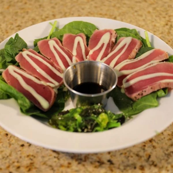 Seared tuna slices with white sauce drizzle, served on a bed of spinach with a side of seaweed salad and soy sauce.